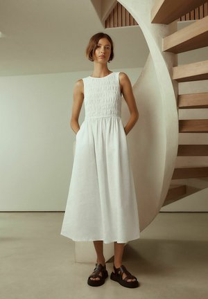 Junge Frau steht mit Händen in den Taschen, trägt ein ärmelloses weißes Midikleid und schwarze Plateausandalen nahe einer geschwungenen Holztreppe.