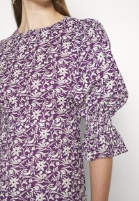 Robe violette avec un motif floral blanc, présentant des manches bouffantes avec des poignets élastiqués et un col rond. Fabriquée en tissu léger.