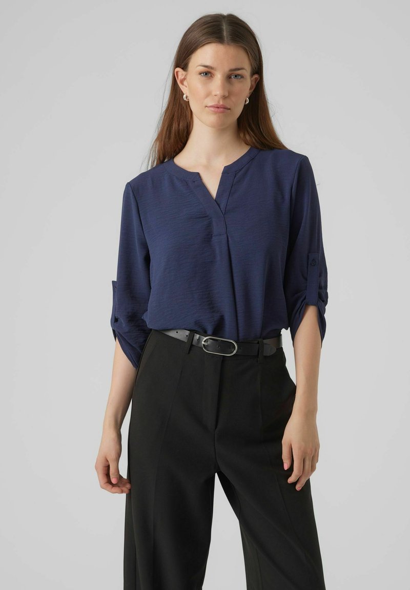 Navyblauwe V-hals blouse met opgerolde driekwartmouwen, gecombineerd met hoge zwarte broek en een zwarte riem met een zilveren gesp.