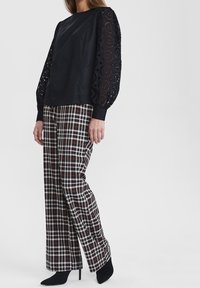 Top nero con maniche a palloncino in pizzo ricamato, abbinato a pantaloni a gamba larga in plaid nei colori nero, bianco e marrone. Scarpe nere con tacco.