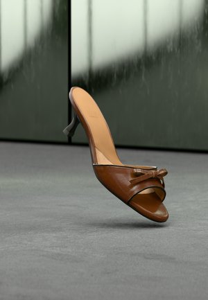 Sandale à talon en cuir marron avec un design croisé et un détail de nœud décoratif. Texture lisse avec un bord noir contrastant, talon surélevé.