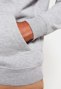 La mano di una persona dentro la tasca anteriore di una felpa grigia chiara con polsini e bordo a coste, indossando pantaloni beige.