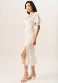 White midi dress with a square neckline, puffed sleeves, ruffled hem, and side slit. Worn with strappy yellow heels. Textured fabric.