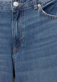 Detailný záber na prednú stranu modrých denimových džínsoch s kovovým gombíkom, zipsom, oranžovým prešívaním a detailmi prednej vrecka.