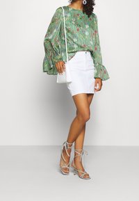 Blusa verde con stampa floreale e maniche a campana, abbinata a una mini gonna di denim bianca. Il modello indossa sandali con tacco a strappo grigio chiaro e tiene una borsa bianca.