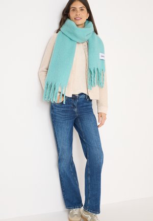 Mujer con una chaqueta texturizada beige, suéter crema, jeans azules, zapatillas beige y una gran bufanda de flecos azul claro, de pie contra una pared blanca.