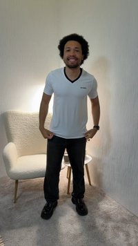 Άνδρας με σγουρά μαλλιά, φορώντας λευκό μπλουζάκι με V-λαιμόκοψη και μαύρο παντελόνι, στέκεται και χαμογελά σε ένα δωμάτιο με υφασμάτινη πολυθρόνα και μικρό ξύλινο τραπέζι.