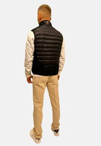 Gilet noir et rembourré avec un col montant, présentant une texture lisse et une poche arrière. Porté sur une chemise blanche à manches longues et un pantalon beige.