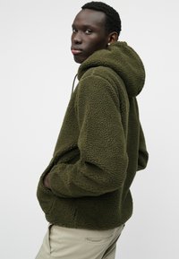 Ελιάς πράσινη φλις ζακέτα με υφή, υπερμεγέθη κουκούλα και πλαϊνές τσέπες, φοριέται πάνω από ανοιχτόχρωμο παντελόνι.