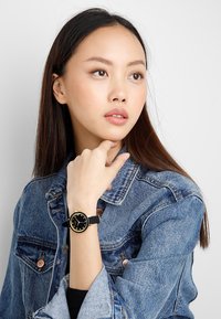 Reloj de pulsera negro con una esfera redonda acentuada en dorado y marcadores de horas simples. Lleva en la muñeca, combinado con una chaqueta de denim.