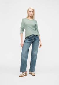 Ξανθιά γυναίκα που στέκεται, φορώντας ένα μακρυμάνικο τοπ σε απόχρωση φασκόμηλου, μπλε τζιν παντελόνι με φαρδιά πόδια και κορδόνια μπροστά, και sneakers με λεοπάρ σχέδιο.