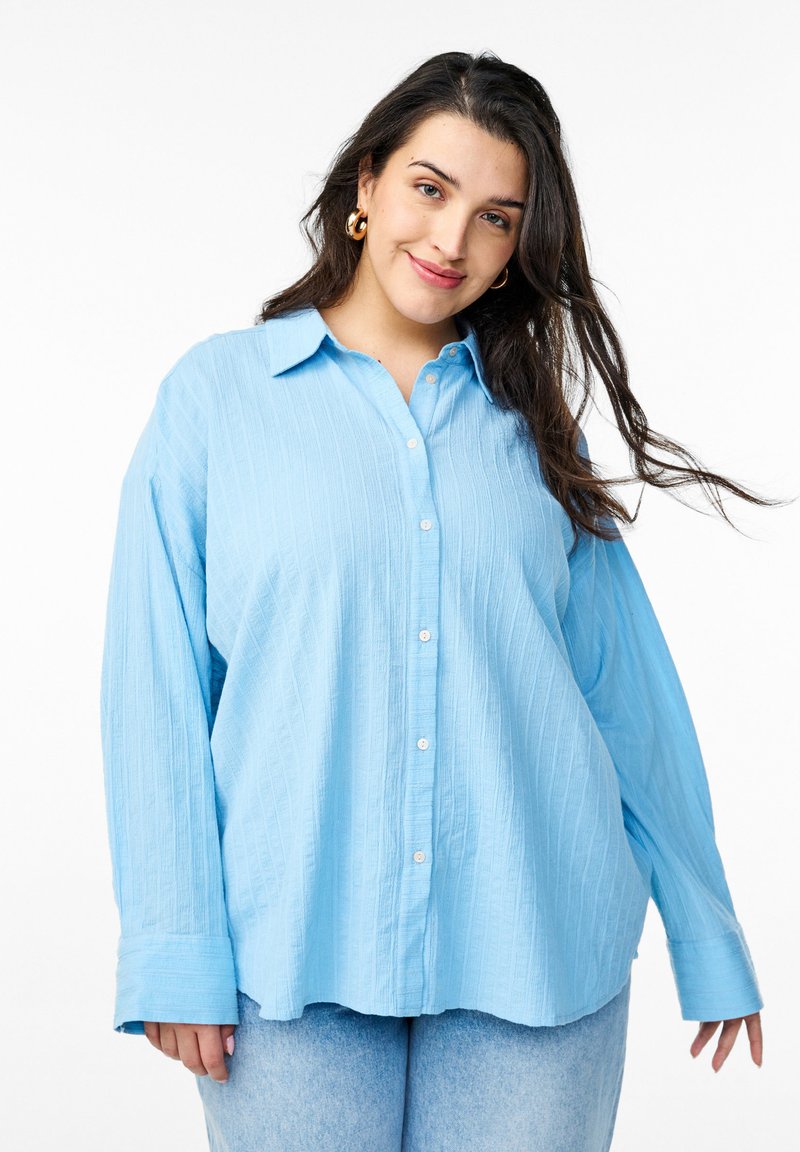 Lys blå skjorte med knapper og tekstureret stof, med krave, lange ærmer og en afslappet pasform. Sammen med lys blå jeans.