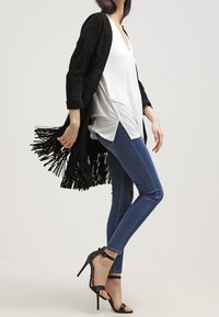Cardigan de ante negro con flecos sobre una blusa de cuello en V blanca, combinado con jeans ajustados azules y sandalias de tacón negro.