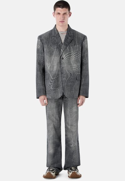 Hombre vistiendo un traje holgado con patrón de cuadros distorsionados en tonos grises, con pantalones a juego y zapatillas, de pie contra un fondo liso.