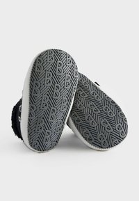 Calzado en blanco y negro con suelas de goma texturizadas que presentan un agarre con patrón y el logotipo 'B' en relieve. Material suave con diseño de punta redonda.