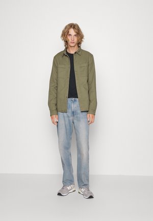 Olivengrønn skjorte med knapper over en svart t-skjorte, sammen med lyseblå jeans og grå sneakers. Rett snitt, uformelt utseende.