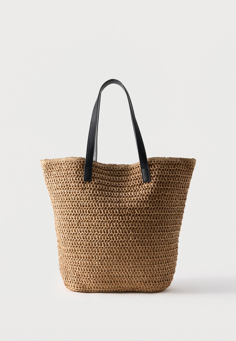 Sac cabas tissé de couleur sable avec des bretelles d'épaule en cuir noir, positionné debout contre un fond clair uni.