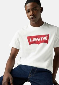 Ung mand iført en hvid Levi's t-shirt med et rødt logo, kombineret med mørkeblå denim jeans, siddende mod en lys grå baggrund.