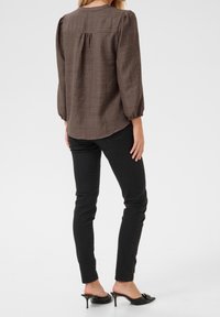 Blusa marrón texturizada con mangas largas y detalles fruncidos en la parte posterior, combinada con vaqueros ajustados negros y mules negros de tacón.