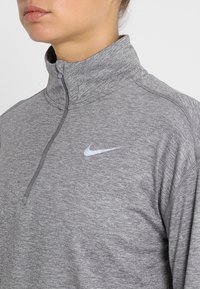 Pullover grigio con zip a quarti realizzato in un tessuto testurizzato. Presenta un colletto alto e un piccolo logo Nike bianco sul lato sinistro del petto.