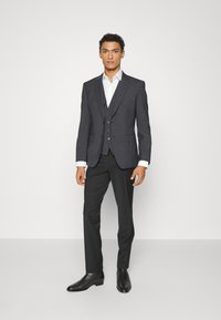 Kolkasgrå skräddarsydd kostym med slagkrage, vit skjorta och svarta dressskor. Den figursydda designen inkluderar en väst med tre knappar.
