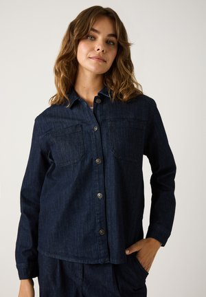 Young woman with wavy brown hair wearing dark blue denim jacket and matching pants, hand in pocket, posing against plain background.