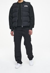 Svart pufferväst med dragkedja fram och texten "PEGADOR" på bröstet, ihop med en svart sweatshirt och cargo-byxor, samt vita sneakers.