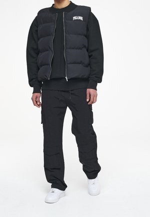 Schwarze Pufferweste mit Reißverschluss und dem Text "PEGADOR" auf der Brust, kombiniert mit einem schwarzen Sweatshirt und Cargohosen sowie weißen Sneakern.
