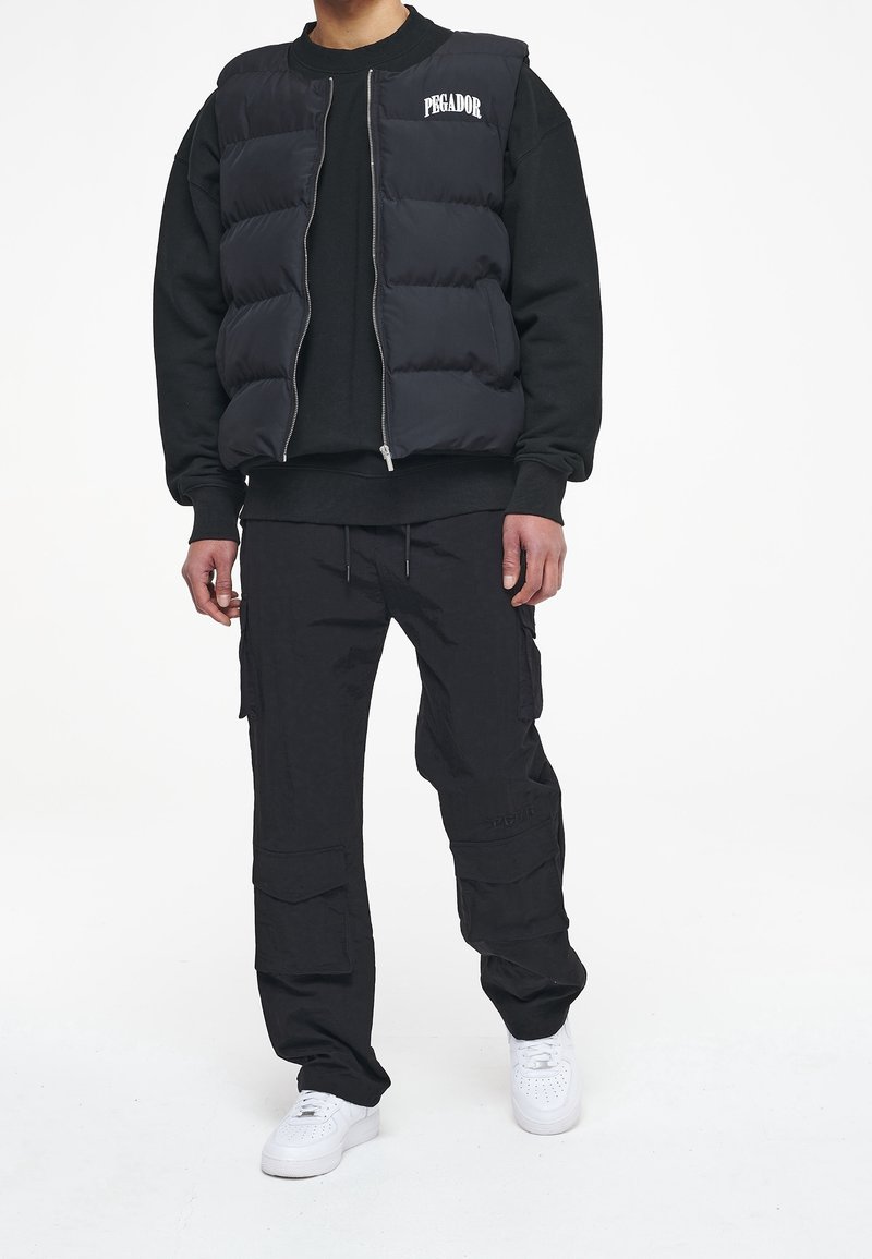 Svart pufferväst med dragkedja fram och texten "PEGADOR" på bröstet, ihop med en svart sweatshirt och cargo-byxor, samt vita sneakers.