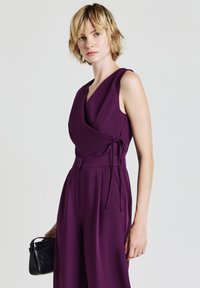 Junge Person mit kurzem blonden Haar, die einen ärmellosen, tiefpurpurnen Overall trägt und eine kleine schwarze Handtasche vor einem schlichten Hintergrund hält.