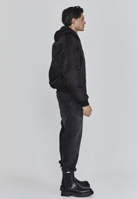 Jonge man met donker haar staat in profiel, gekleed in een zwarte hoodie, zwarte jeans en zwarte stevige laarzen tegen een effen grijze achtergrond.