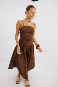 Smeđa haljina bez rukava koja ima uski gornji dio i lepršavu, nabranju suknju. Model nosi ravne, pletene sandale i razne narukvice.