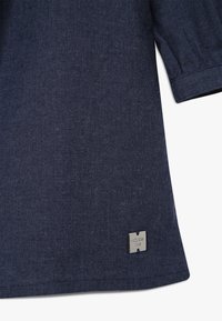 Ciemnoniebieska tkanina z krótkim rękawem i małą srebrną metalową metką w kształcie kwadratu blisko dolnego brzegu na białym tle.