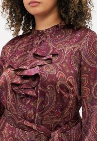 Blusa viola a paisley con dettagli arricciati, chiusura con bottoni e una cintura. Materiale liscio con dettagli intricati in verde e rosso.