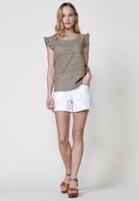 Top de crochet en caqui claro con mangas cortas acampanadas, combinado con pantalones cortos blancos. La modelo lleva sandalias de plataforma marrones. Fondo liso.