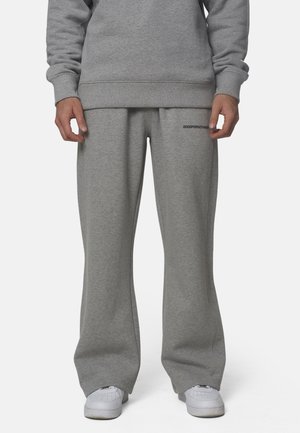 Onderlichaam van een persoon die losse grijze joggingbroek draagt met een lichte uitlopende pijp en witte sneakers, handen ontspannen langs de zijkant.