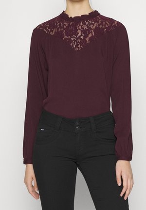 Blouse bordeaux à manches longues avec un délicat panneau en dentelle au niveau du col, assortie à un pantalon noir ajusté avec une fermeture à double bouton.