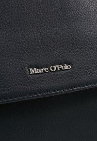 Surface en cuir noir texturé avec logo de la marque "Marc O'Polo" en argent et couture visible le long du bord.