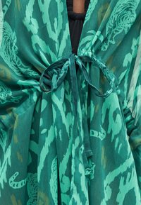 Πράσινο ύφασμα με σχέδια δεμένο με φιόγκο μπροστά πάνω από μαύρο ένδυμα, που δείχνει υφή και πτυχές στο ελαφρύ υλικό.