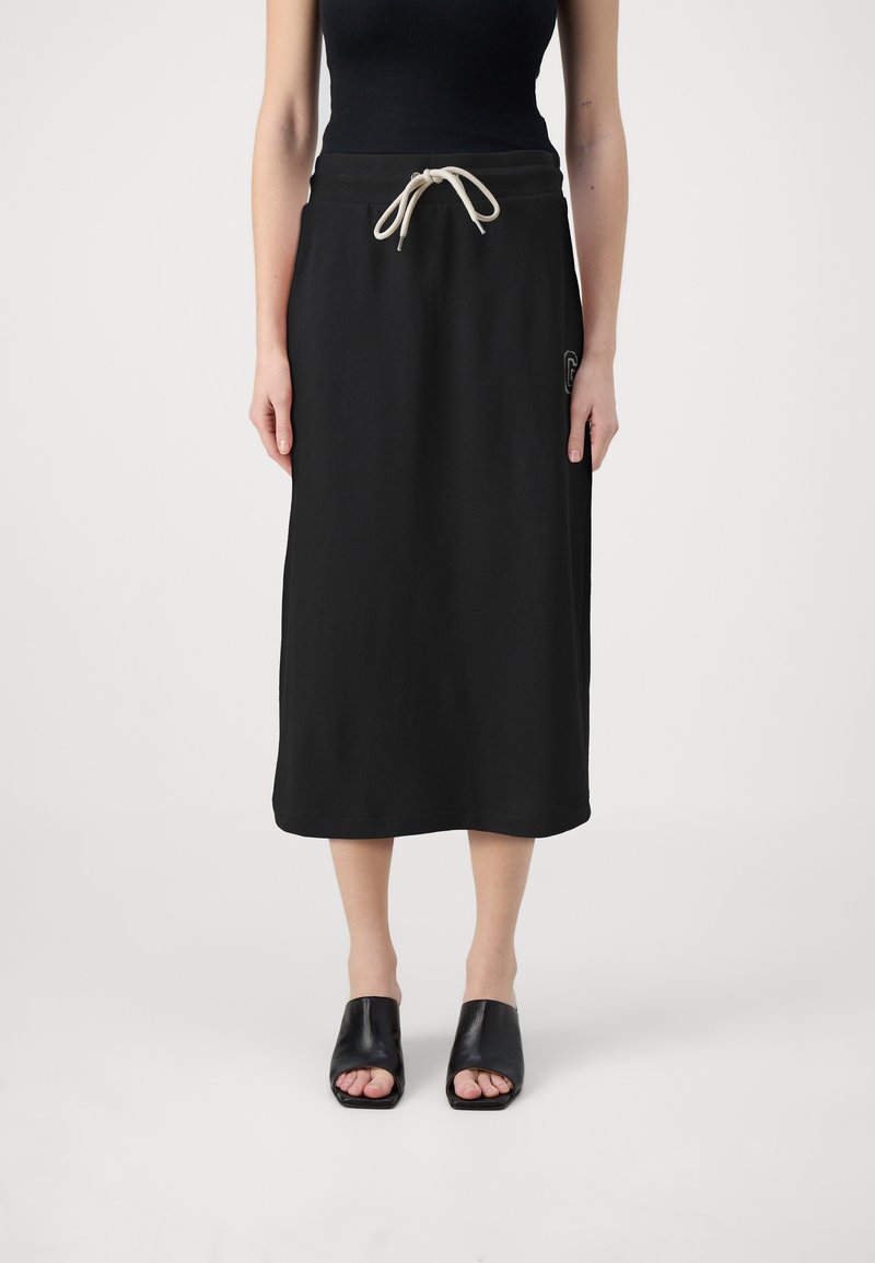 Femme portant une jupe midi noire avec un cordon blanc, un haut sans manches noir et des mules noires à bout ouvert, debout sur un fond blanc.