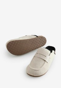 Beige Filzslipper mit einem Ledersteifen, weicher schwarzer Innensohle und einer strukturierten Gummisohle mit geometrischem Muster.
