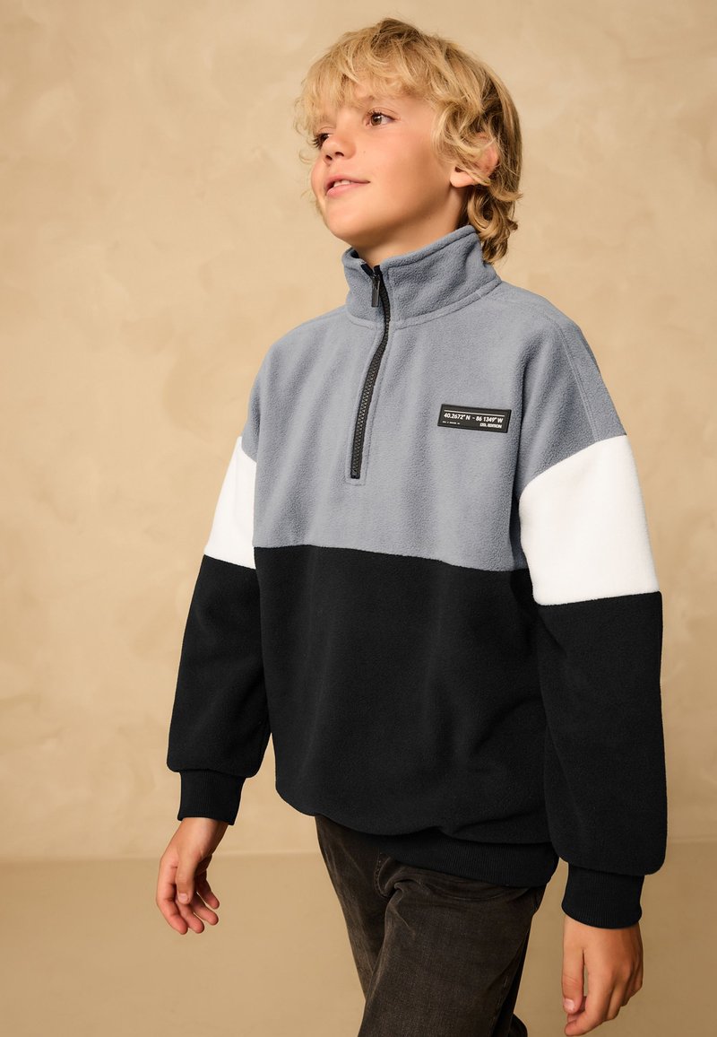Garçon blond portant un pull polaire bicolore avec des sections grises, blanches et noires, debout devant un fond beige.