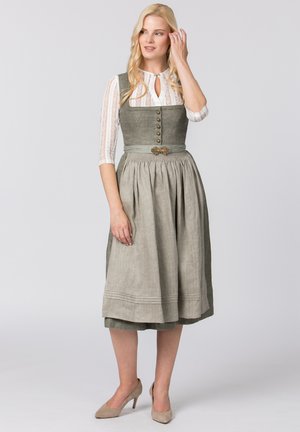 Blonde Frau trägt ein graues traditionelles Kleid mit geknöpftem Mieder, weißer Spitzenbluse, Schürze und beigen High Heels, steht vor einem einfarbigen Hintergrund.