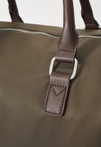 Brown leather strap with metal hardware attached to an olive green fabric bag. The texture is smooth with visible stitching details.