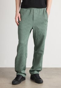 Grüne Sweatpants mit einem strukturierten geometrischen Design, elastischem Bund und Seitentaschen, kombiniert mit schwarzen Sneakers.