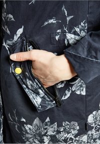 Námořnická bunda s květinovým vzorem a kapsou na zip. Tkanina je hladká, s bílými květinami na tmavém pozadí a detailem na patentce.