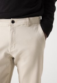 Homme portant un pantalon beige et un pull noir à manches longues, la main droite dans la poche, sur un fond uni.