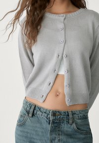 Light gray knitted cardigan with long sleeves, featuring six lavender buttons and ribbed hem. Paired with mid-rise, faded blue jeans.