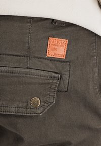 Pantaloni verzi măslinii cu un finisaj texturat, prezentând o aplicație cu logo maro și un buton din alamă pe buzunar, concepuți pentru funcționalitate.