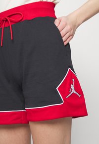Pantalones cortos negros con una cinturilla acanalada roja, que presentan detalles en blanco y un logo gris de Jordan en la parte inferior izquierda. Cuerda ajustable para mayor comodidad.